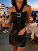 A woman at an outdoor evening restaurant wears a black halter top with a large bow detail and a rhinestone-embellished square center, paired with a black mini skirt. She holds a glass of champagne, with tables, dining chairs, and a sunset seascape in the background.