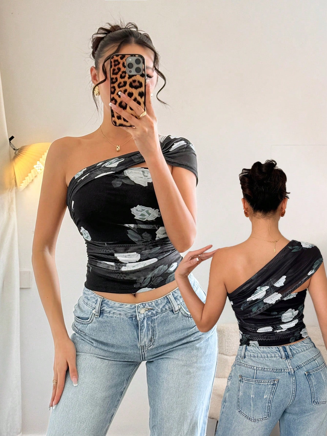 Woman wearing a black one-shoulder top with white floral patterns and blue jeans, taking a mirror selfie.