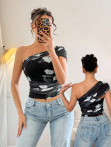 Woman wearing a black one-shoulder top with white floral patterns and blue jeans, taking a mirror selfie.