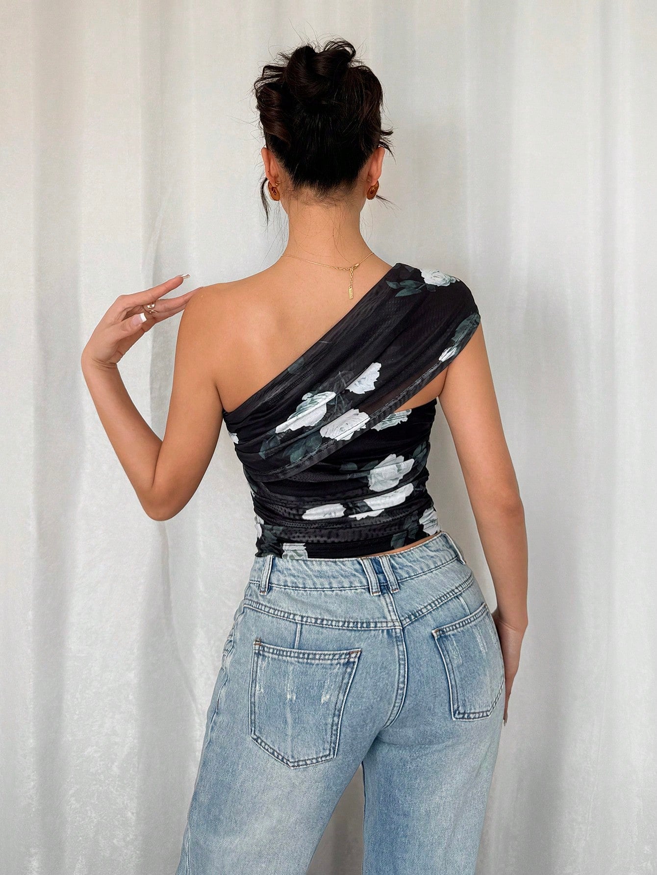 Person wearing a black one-shoulder top with white patterns and light blue jeans against a white curtain background.