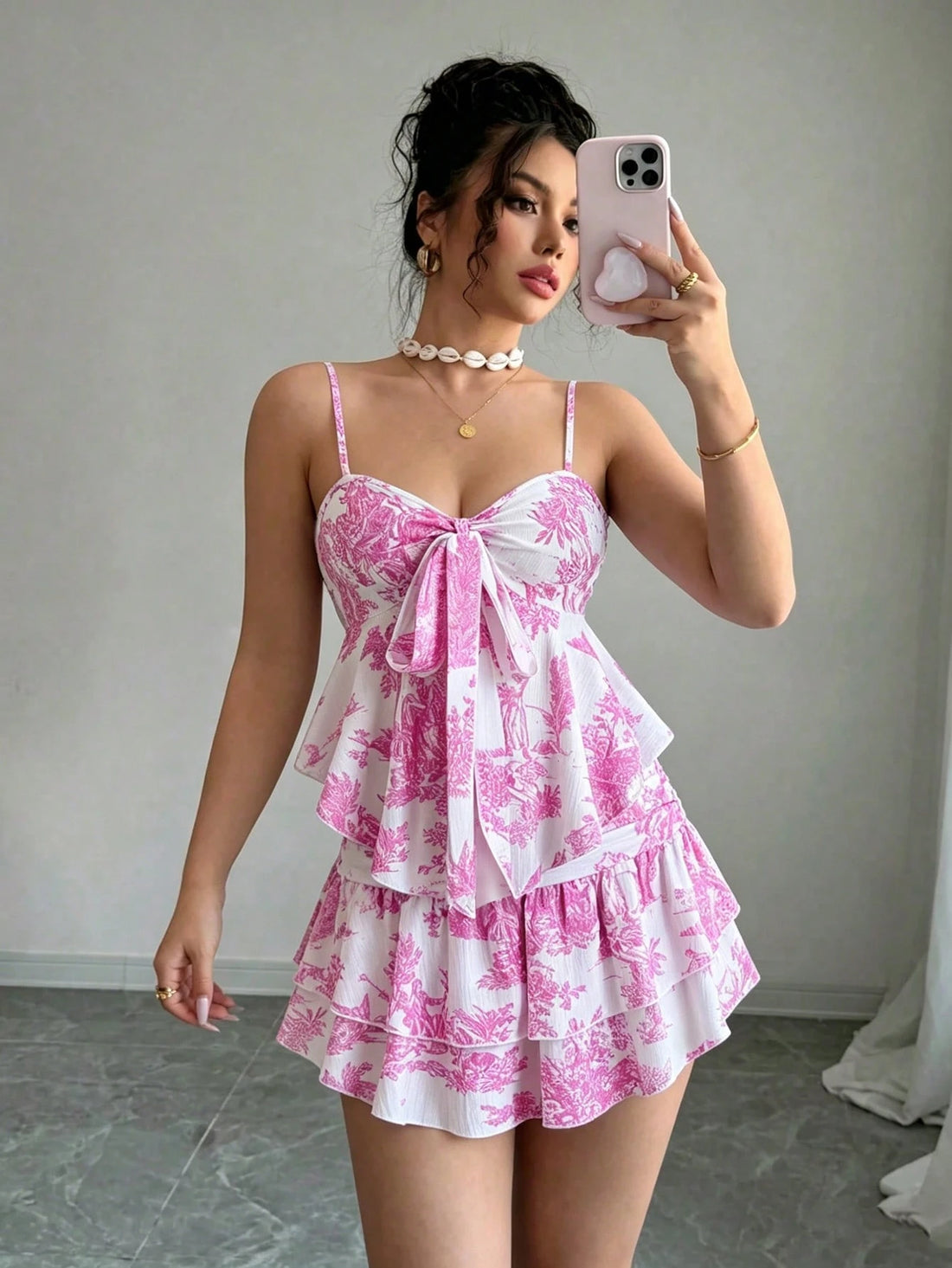 A woman takes a mirror selfie wearing a pink and white toile-patterned mini dress with spaghetti straps, a large bow at the bust, and a ruffled tiered skirt.