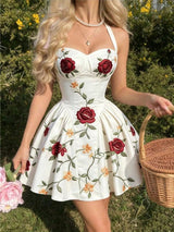 A woman wearing a white halter mini dress with red rose and yellow floral embroidery, holding a wicker basket and pink flowers in an outdoor garden setting.