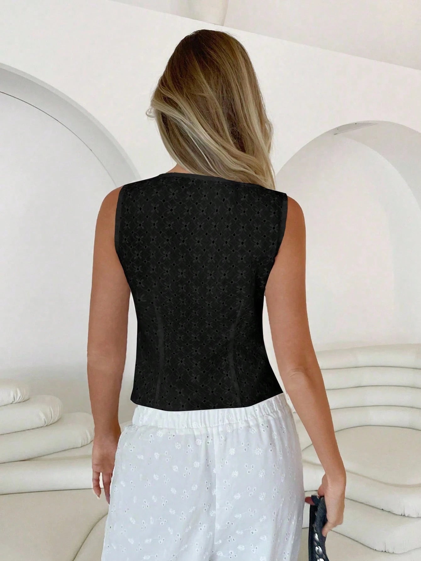 Woman wearing a black sleeveless top and white pants in a white room.