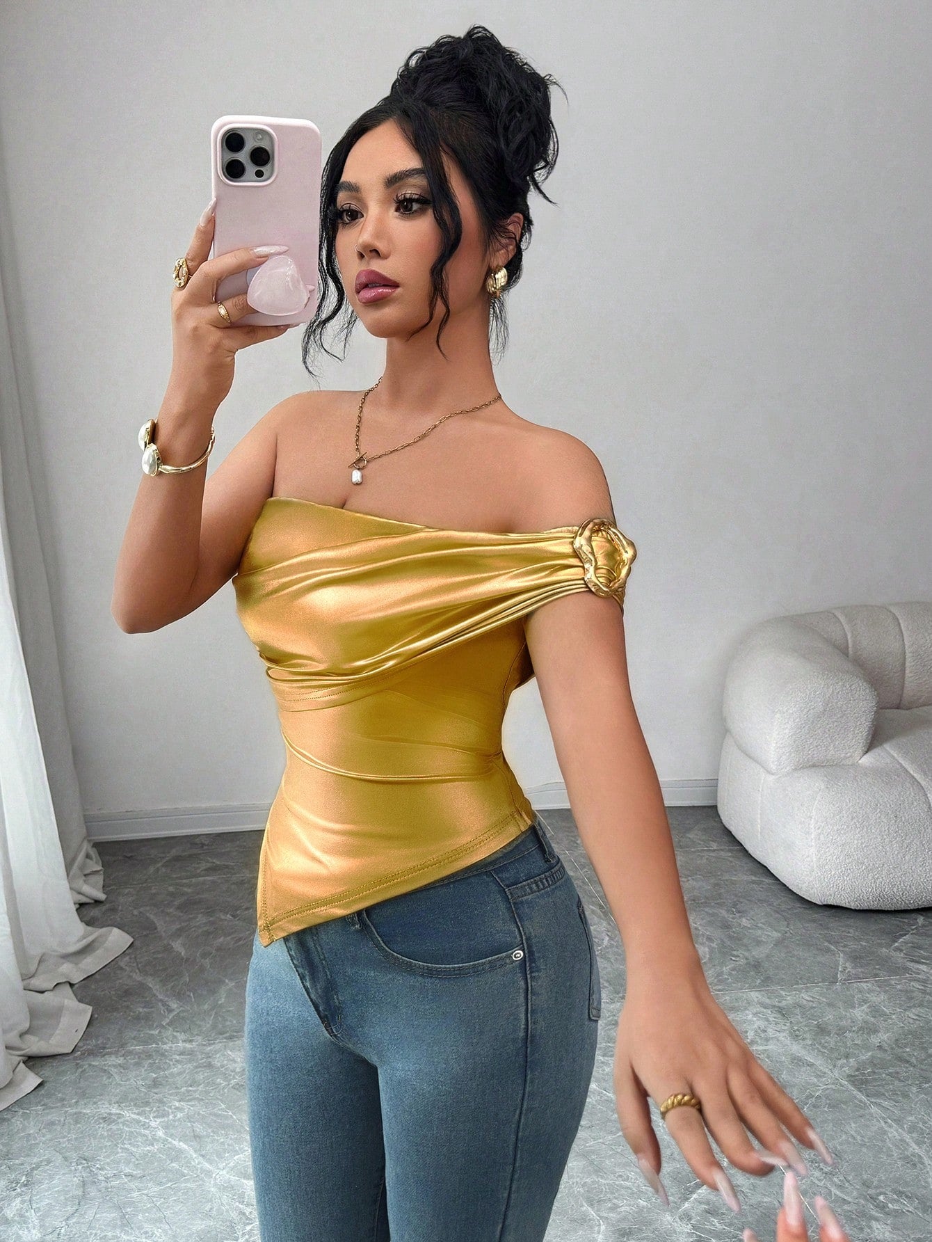 Woman wearing a gold top and blue jeans, taking a mirror selfie.