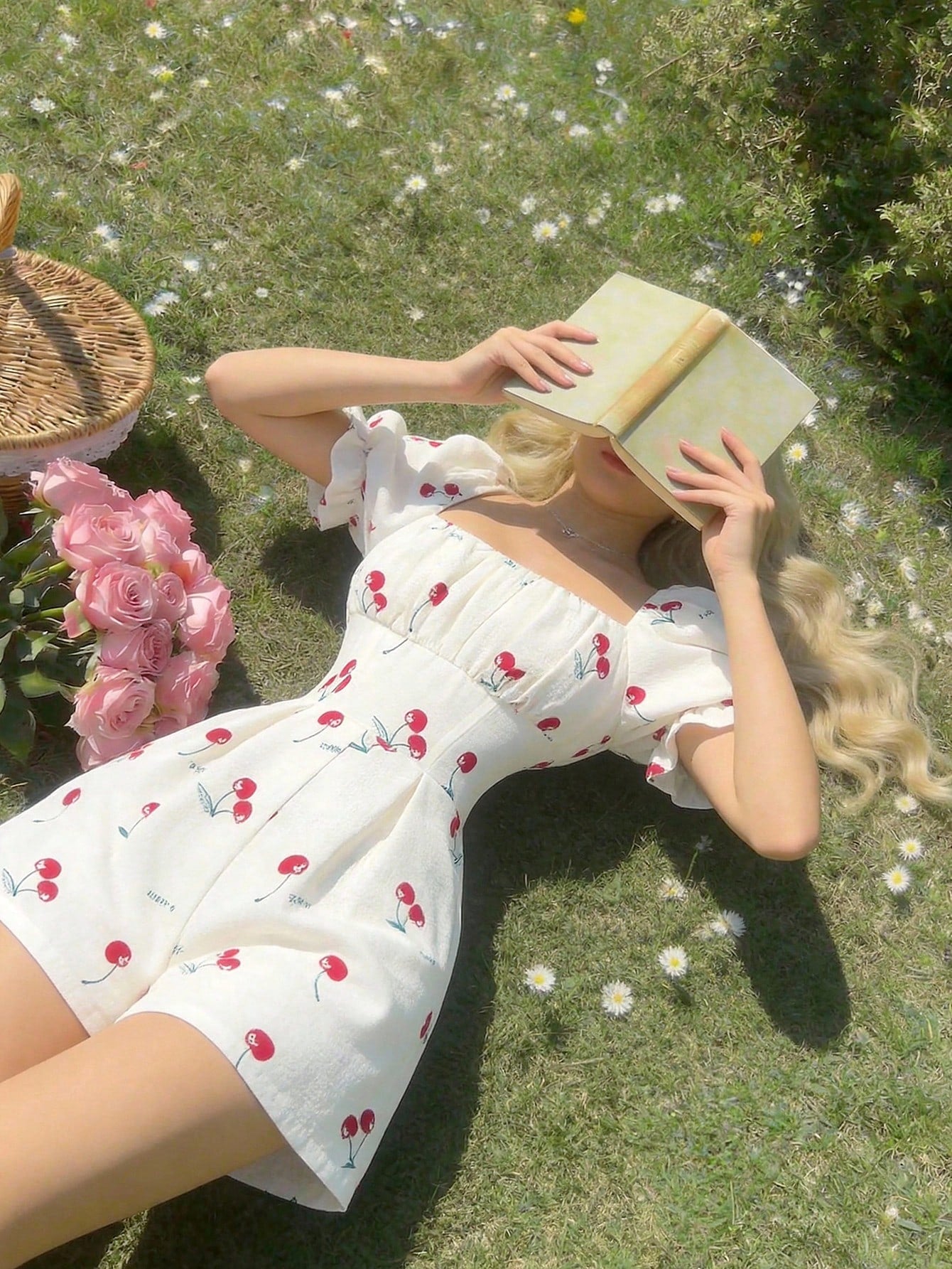 A woman lying on a grassy lawn with wildflowers, wearing a white short romper printed with red cherries. She is holding a book over her face, with pink flowers and a wicker basket nearby.