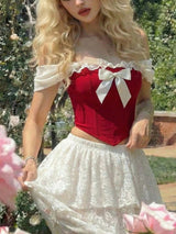 A woman wearing a red off-the-shoulder corset-style top with a large white bow and a white lace skirt, photographed outdoors among greenery and pink flowers.