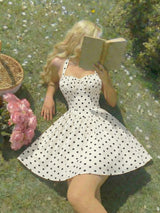 A woman lying on grass outdoors, reading a book. She is wearing a white sleeveless dress with black polka dots.