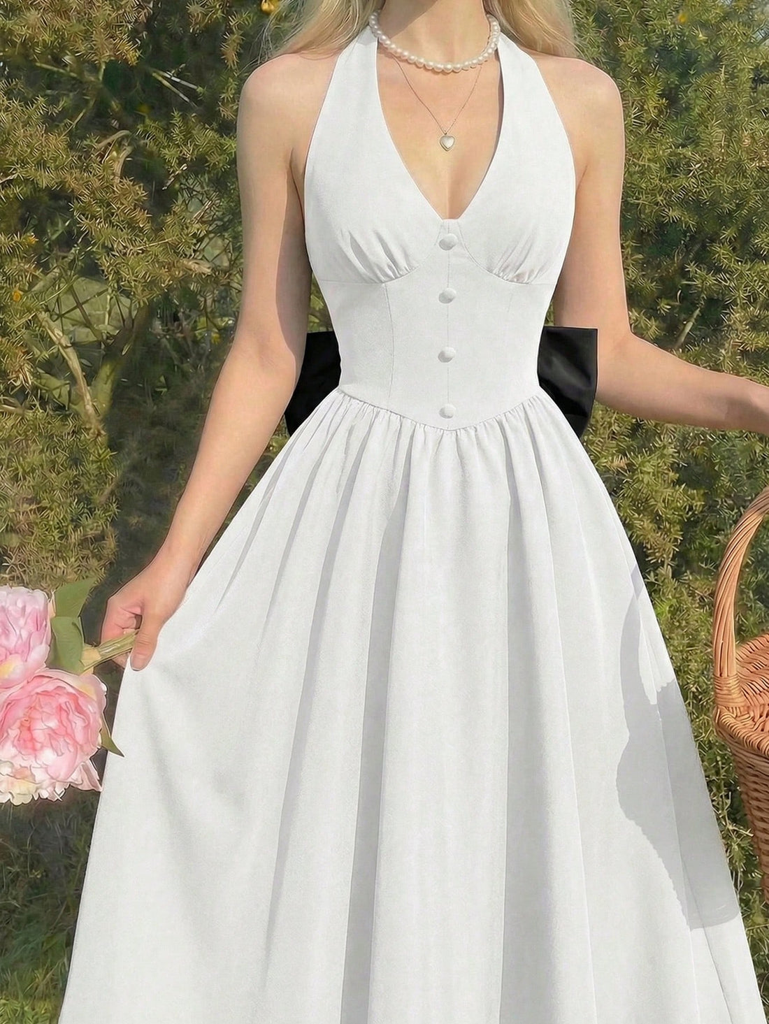 A woman wearing a white halter-neck A-line dress with a buttoned bodice and a black ribbon detail at the waist, standing outdoors in front of green foliage.