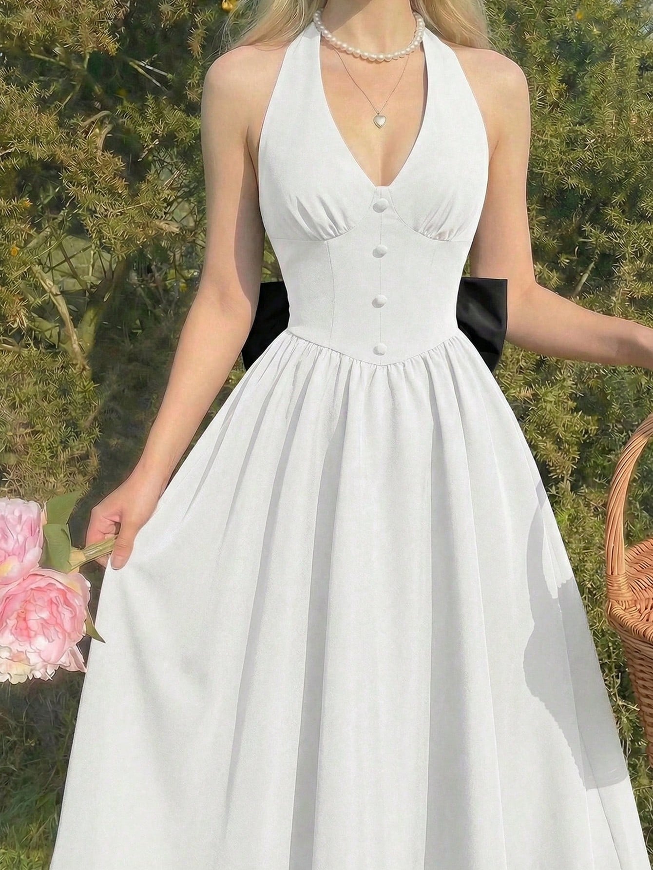 A woman wearing a white halter-neck A-line dress with a buttoned bodice and a black ribbon detail at the waist, standing outdoors in front of green foliage.