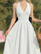 A woman wearing a white halter-neck A-line dress with a buttoned bodice and a black ribbon detail at the waist, standing outdoors in front of green foliage.
