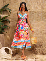 A woman models a vibrant, multicolored floral and geometric print midi dress in a tropical-themed setting with a woven straw backdrop, potted plants, and a straw bag.