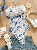 White ruffled bustier bodysuit with a blue floral print, styled in a flat lay with denim shorts, white sandals, a cream handbag, a pearl necklace, and tropical flowers on a beige fabric background.