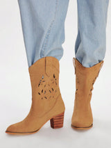 Tan suede western-style ankle boots with laser-cut floral details and stacked wood-look block heels, shown on a model wearing light-wash blue jeans against a white studio background.