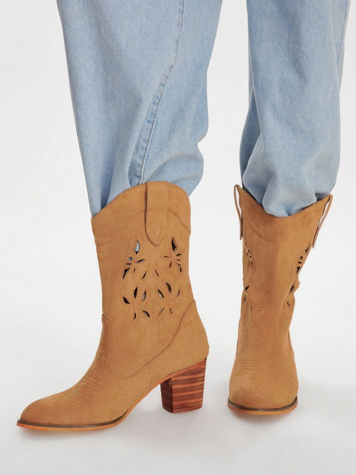 Tan suede western-style ankle boots with laser-cut floral details and stacked wood-look block heels, shown on a model wearing light-wash blue jeans against a white studio background.