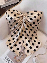 A strapless satin corset-style top with a large gathered bow at the bust and black polka dots on a warm beige background, styled on a light upholstered chair with a pearl necklace and magazine nearby.