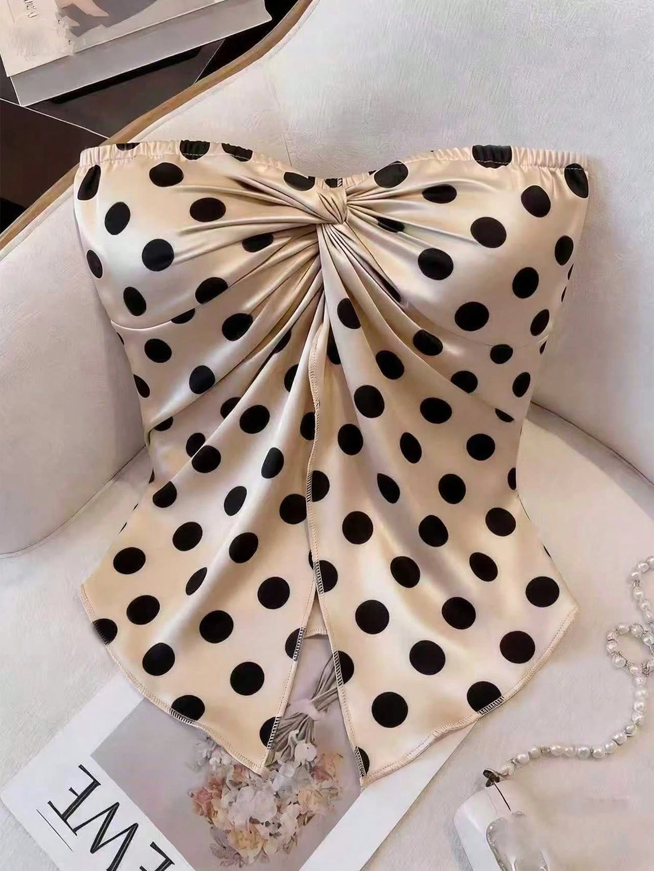 A strapless satin corset-style top with a large gathered bow at the bust and black polka dots on a warm beige background, styled on a light upholstered chair with a pearl necklace and magazine nearby.