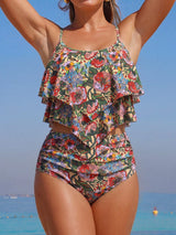 A woman wearing a two-piece ruffled bikini with a colorful floral print on a green base, photographed outdoors against a blue sky and ocean background.