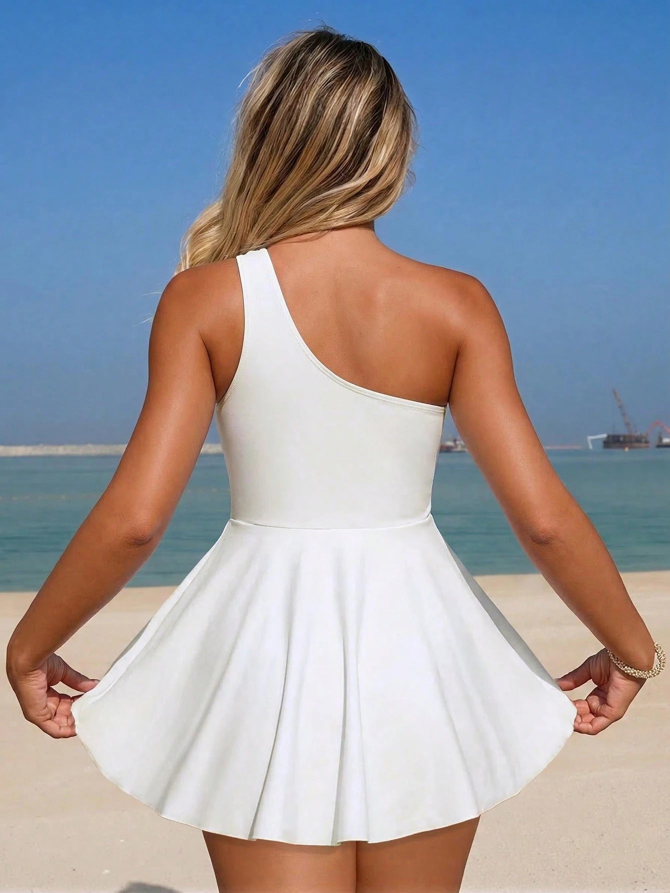 Woman in a white dress standing on a beach with clear blue sky and ocean.