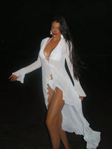 A woman posing in a flowing white sheer long-sleeve cover-up dress against a solid black background.