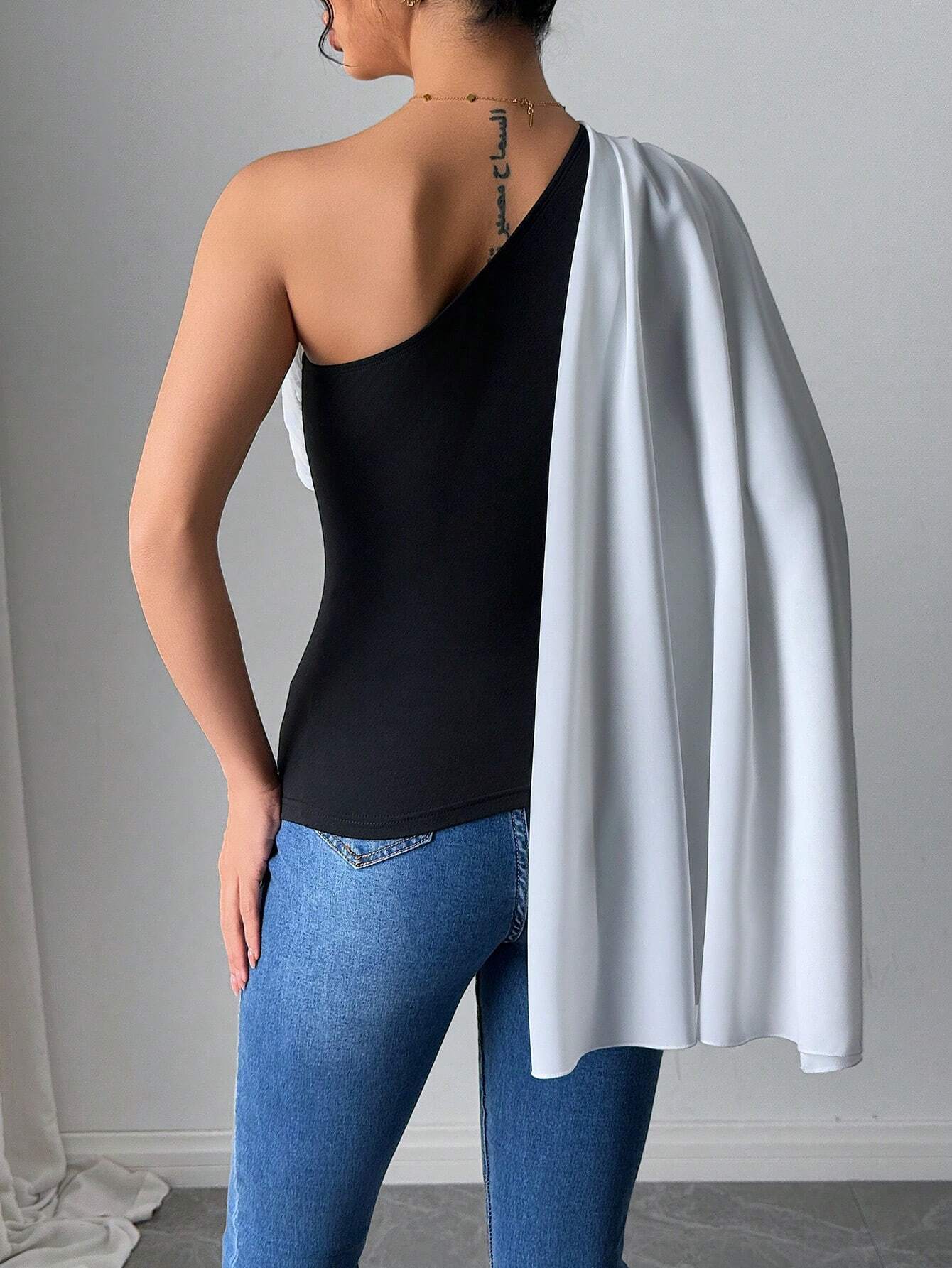 Person wearing a black one-shoulder top, blue jeans, and a white cape over their shoulder against a gray background.