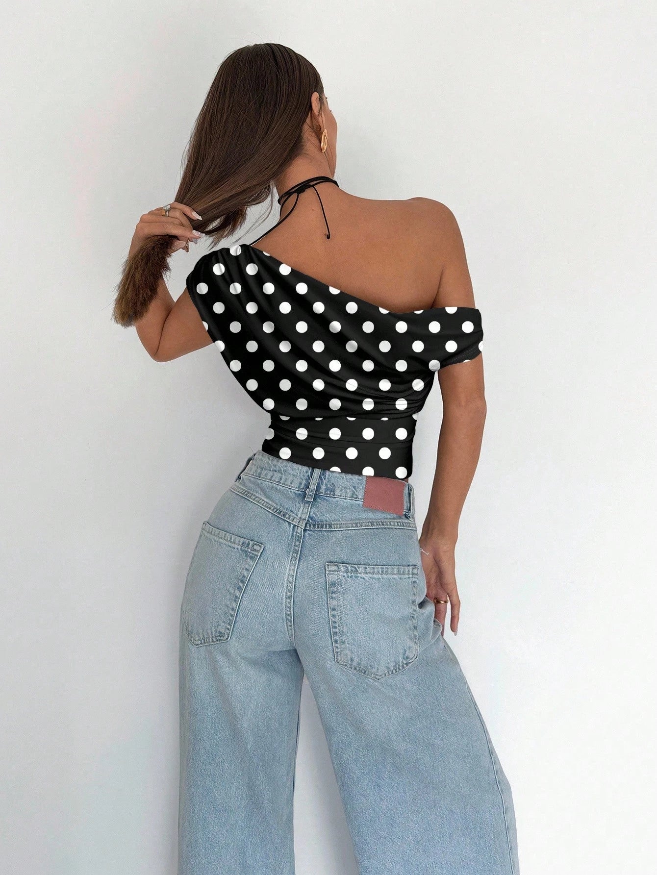 Person wearing a black one-shoulder top with white polka dots and light blue jeans against a white background