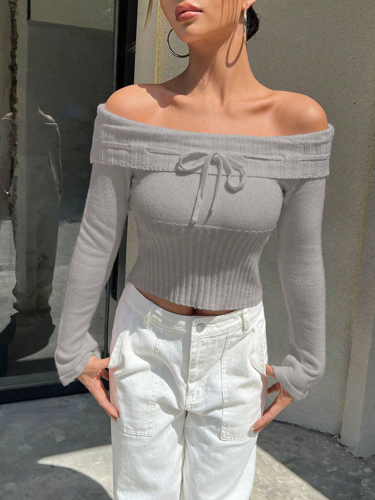 A woman wearing a light gray off-the-shoulder ribbed long-sleeve crop top with a front tie detail, paired with white pants, standing outdoors near a glass door and concrete wall.