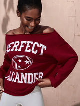 Woman wearing a red sweatshirt with 'PERFECT' and 'ALEXANDER' text against a beige wall.
