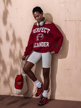 Person wearing a red sweatshirt with text, white shorts, and red sneakers against a beige wall.
