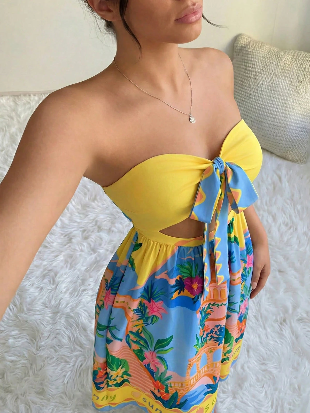A woman wearing a strapless yellow bikini top with a large side tie and a colorful tropical print skirt, standing on a white textured rug.