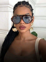Woman wearing large sunglasses and gold earrings against a white wall.