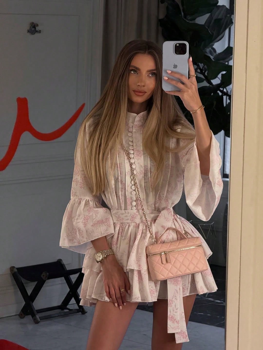 A woman takes a mirror selfie wearing a light pink and white floral print mini dress with long bell sleeves and a ruffled waist. She is holding a light pink quilted crossbody bag with a chain strap and holding a white smartphone.