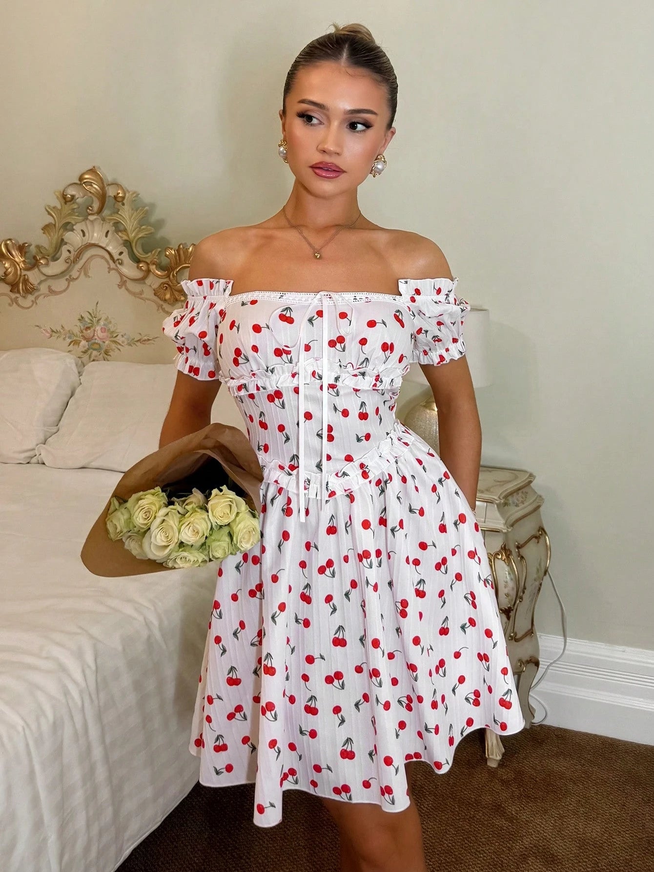 A woman models an off-the-shoulder white dress with a red and black cherry print, holding a brown hat with white roses in a bedroom setting.