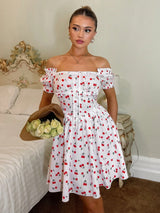A woman models an off-the-shoulder white dress with a red and black cherry print, holding a brown hat with white roses in a bedroom setting.