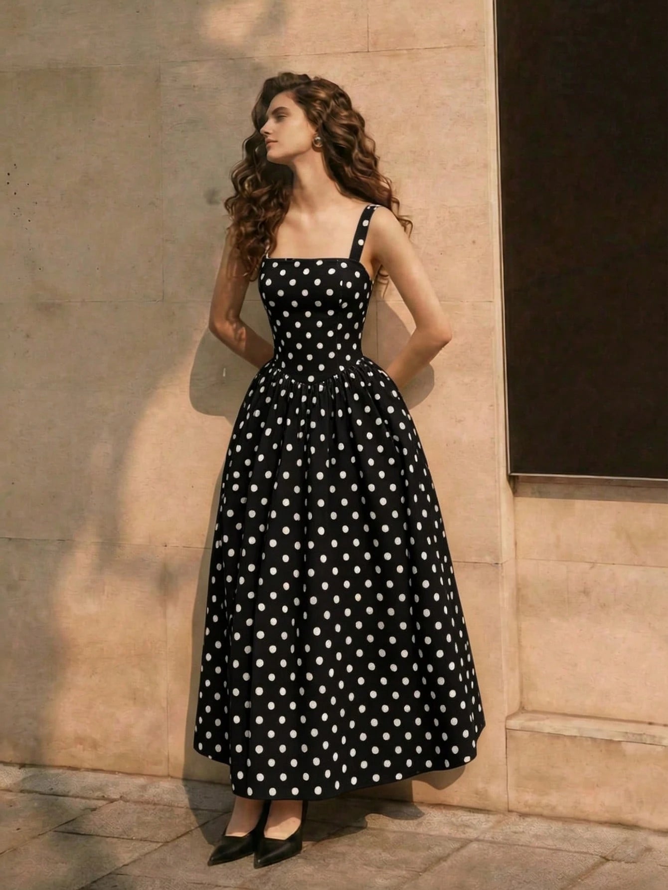 Woman wearing a black dress with white polka dots standing against a beige wall.