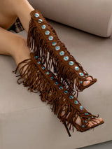 A pair of brown suede-like fringed high-heeled sandals worn on feet, decorated with round turquoise-blue and silver rhinestone embellishments, shown on a beige sofa.