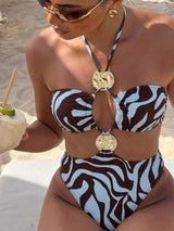 A woman on a beach wearing a brown and white zebra-print bikini with large gold coin pendant details on the halter strap. She is holding a coconut drink and wearing gold sunglasses and earrings.