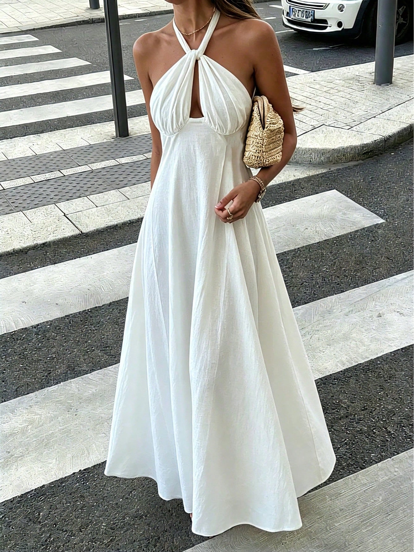 A woman wearing a white halter-neck maxi dress with a gathered bodice and flowing skirt, standing on a city crosswalk and holding a woven straw bag.