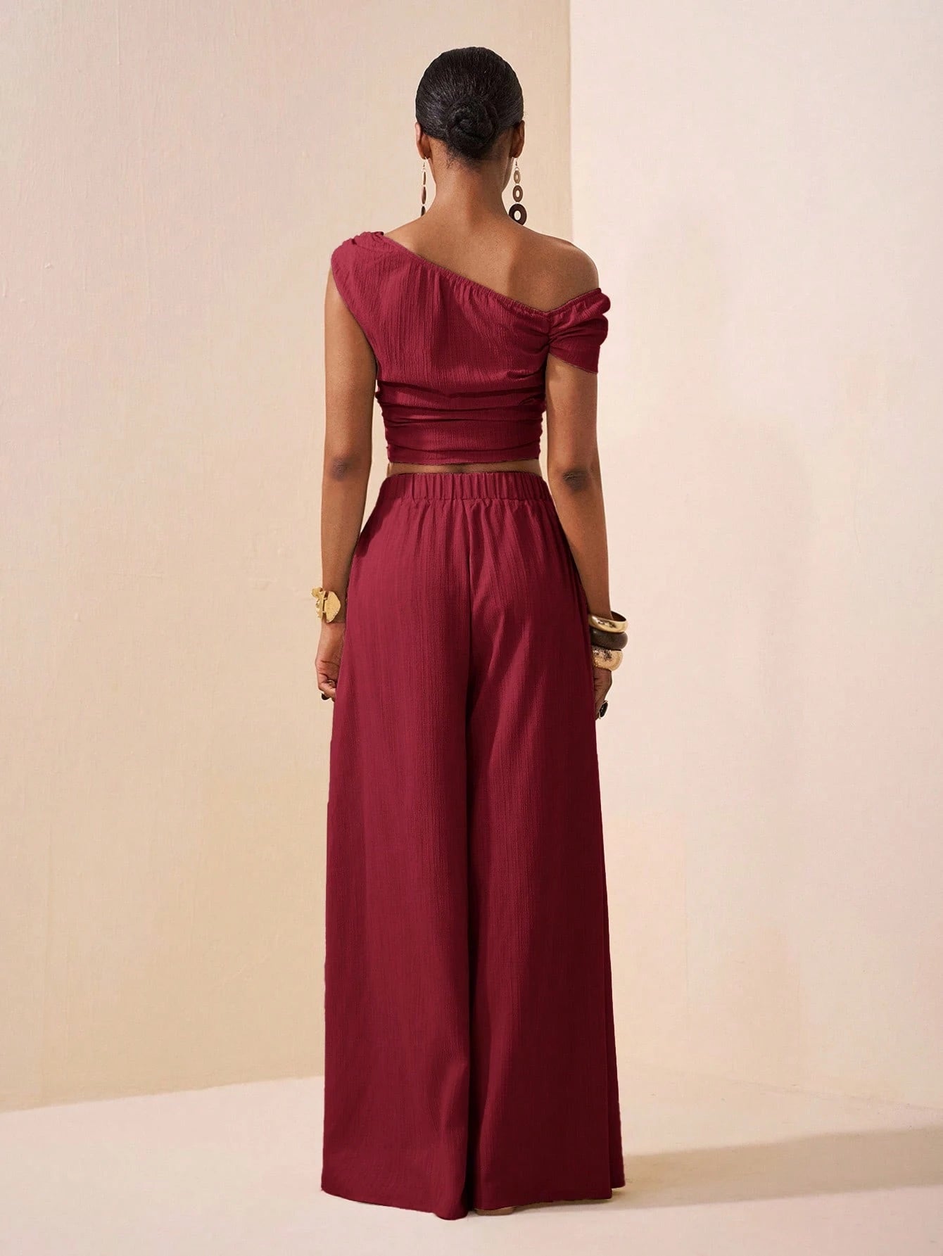 Person wearing a burgundy one-shoulder top and wide-leg pants against a beige background